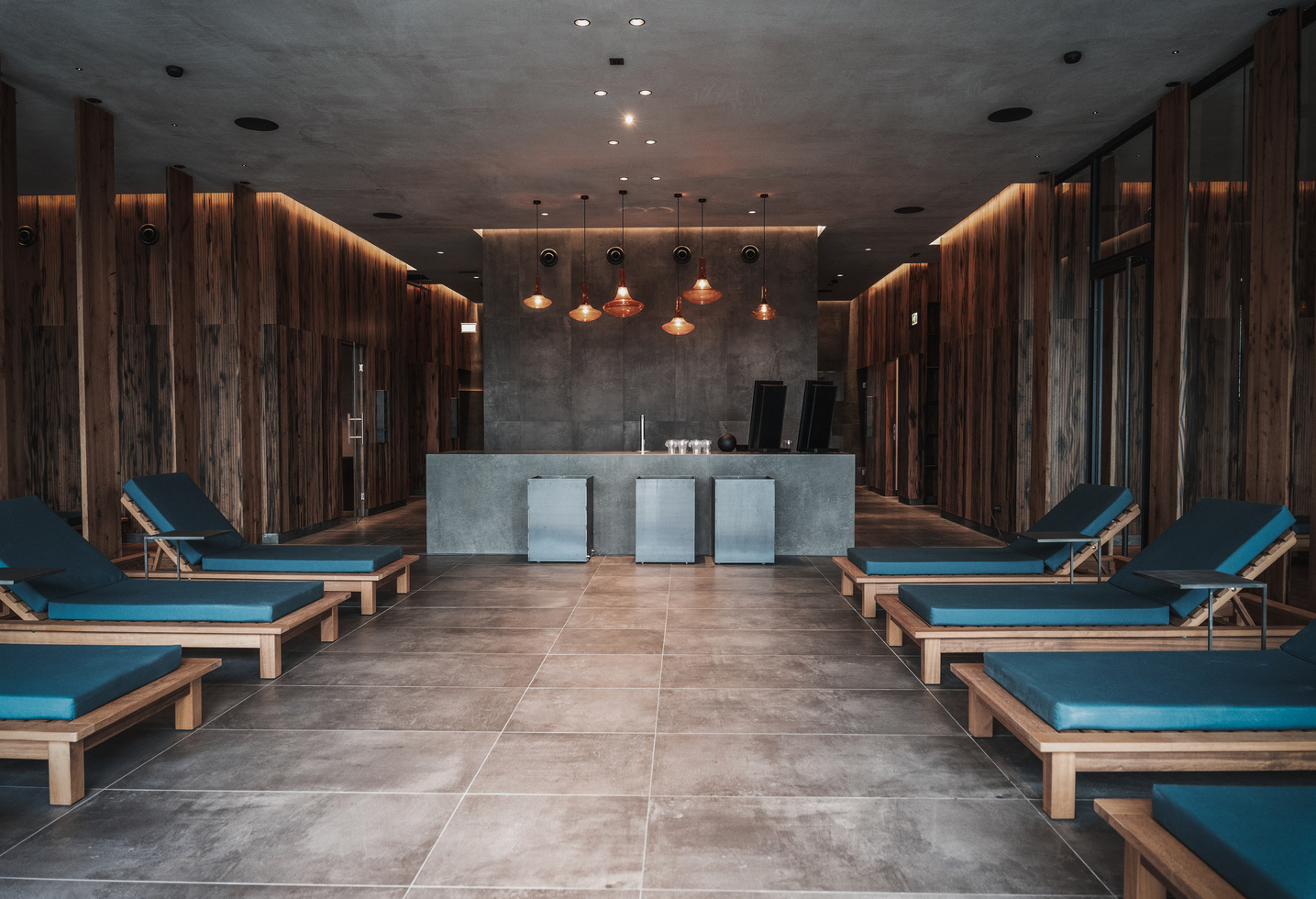 Moderne Spa-Lounge mit hölzernen Chaiselounges und blauen Kissen, gegenüber einem eleganten Empfangstresen mit warmer Beleuchtung.