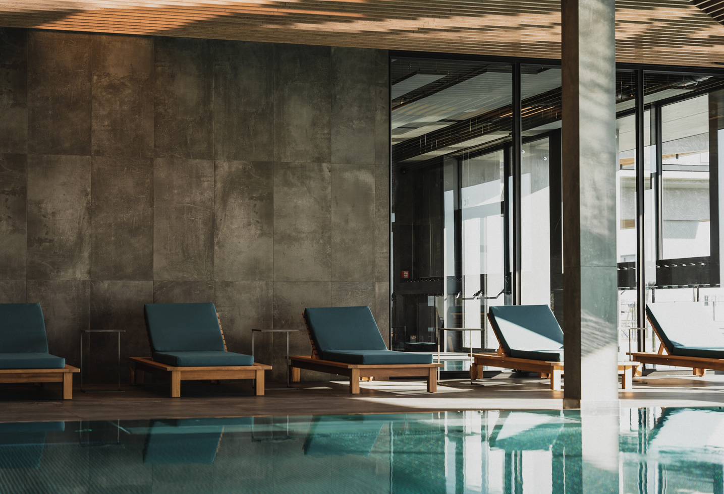 Indoor pool area with blue sun loungers, sunlight streaming through large windows and modern furnishings.