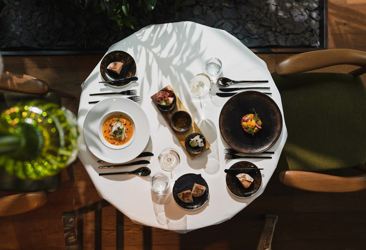 A round table for two with gourmet dishes, bread, drinks and elegant crockery, seen from above.