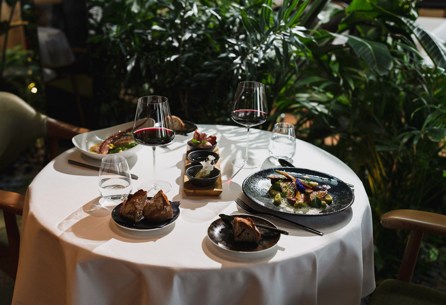 Elegantly laid table for two with wine, bread and gourmet dishes, surrounded by lush green plants.