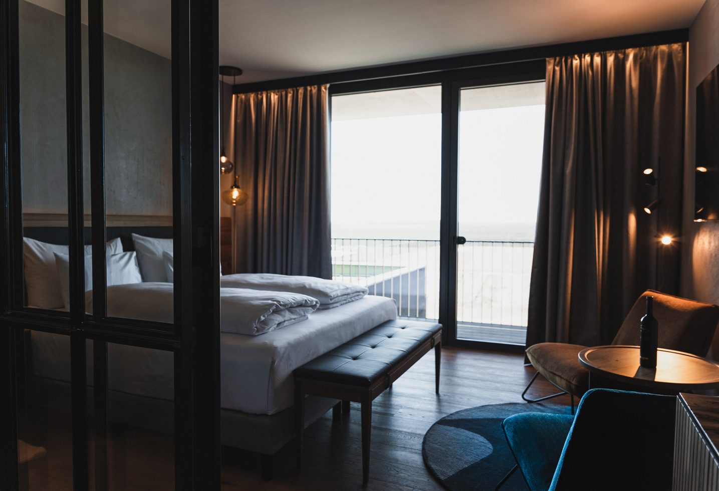 Modern hotel room with large bed, sitting area and floor-to-ceiling windows with curtains.