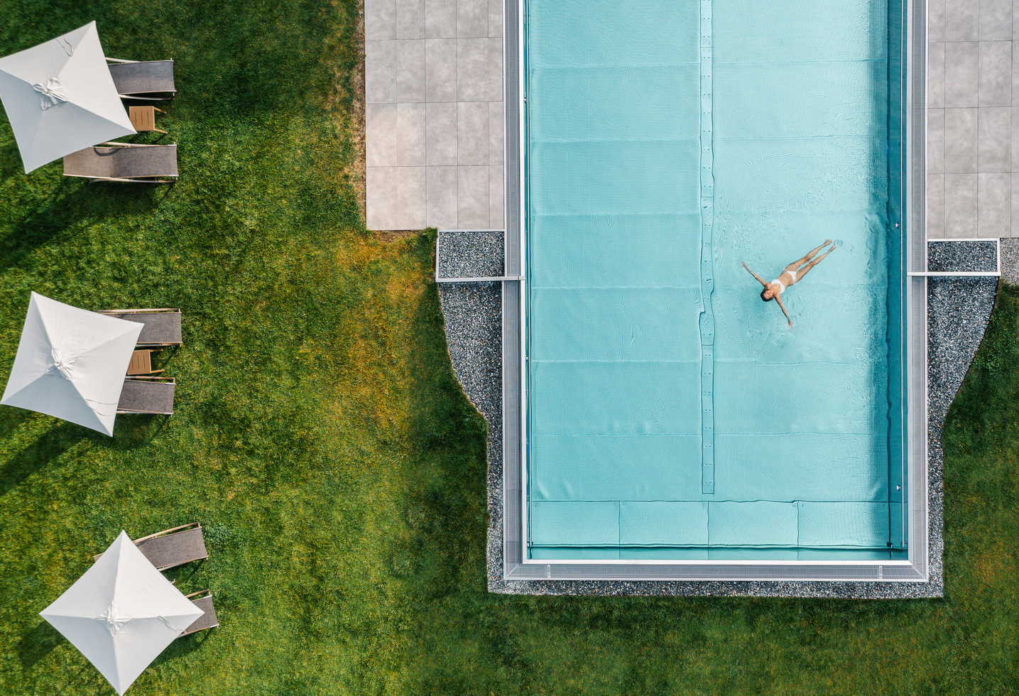 Eine Person schwimmt allein in einem rechteckigen Becken neben grünem Gras und drei Liegestühlen mit weißen Schirmen.