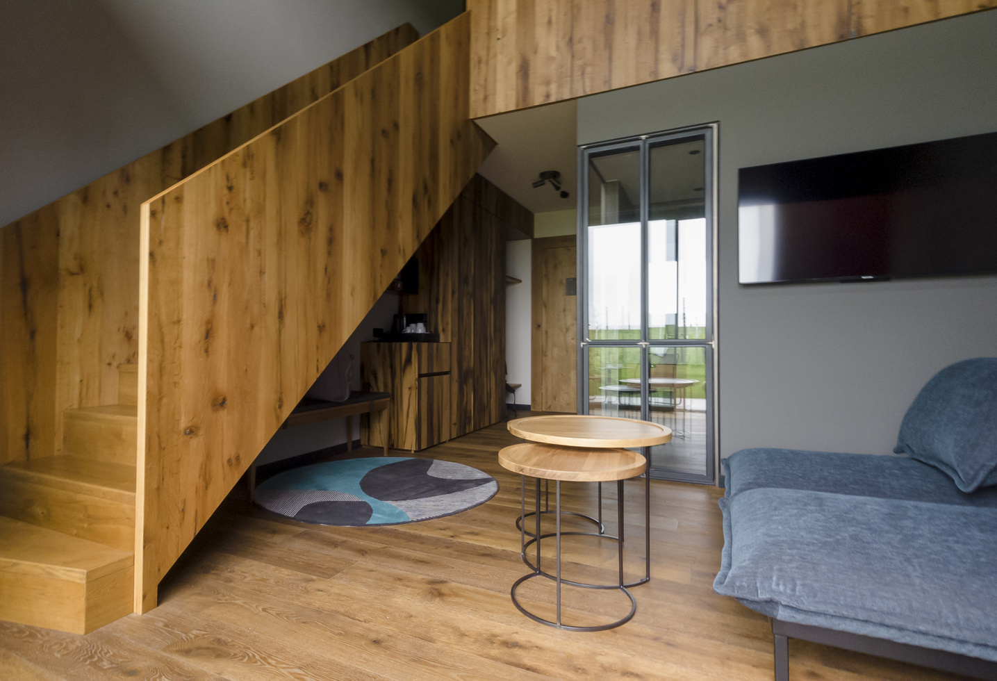 Modern living room with wooden staircase, blue-grey sofa, round tables and wall-mounted TV.
