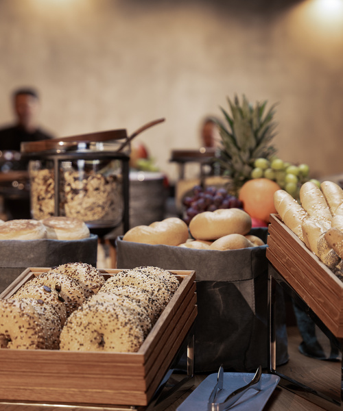 Various bagels and rolls with fruit on a buffet table, blurred people in the background.