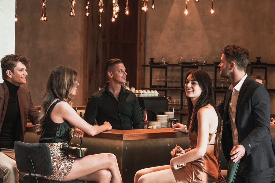 Five stylishly dressed people sit and stand around a bar, smiling and chatting under hanging lamps.
