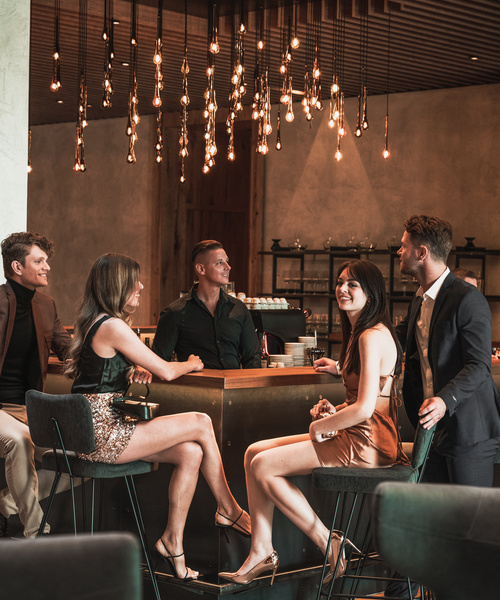 Five stylishly dressed people sit and stand around a bar, smiling and chatting under hanging lamps.