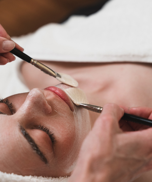 Die Person erhält eine Gesichtsbehandlung, während zwei Bürsten eine Crememaske auf Kinn und Untergesicht auftragen.