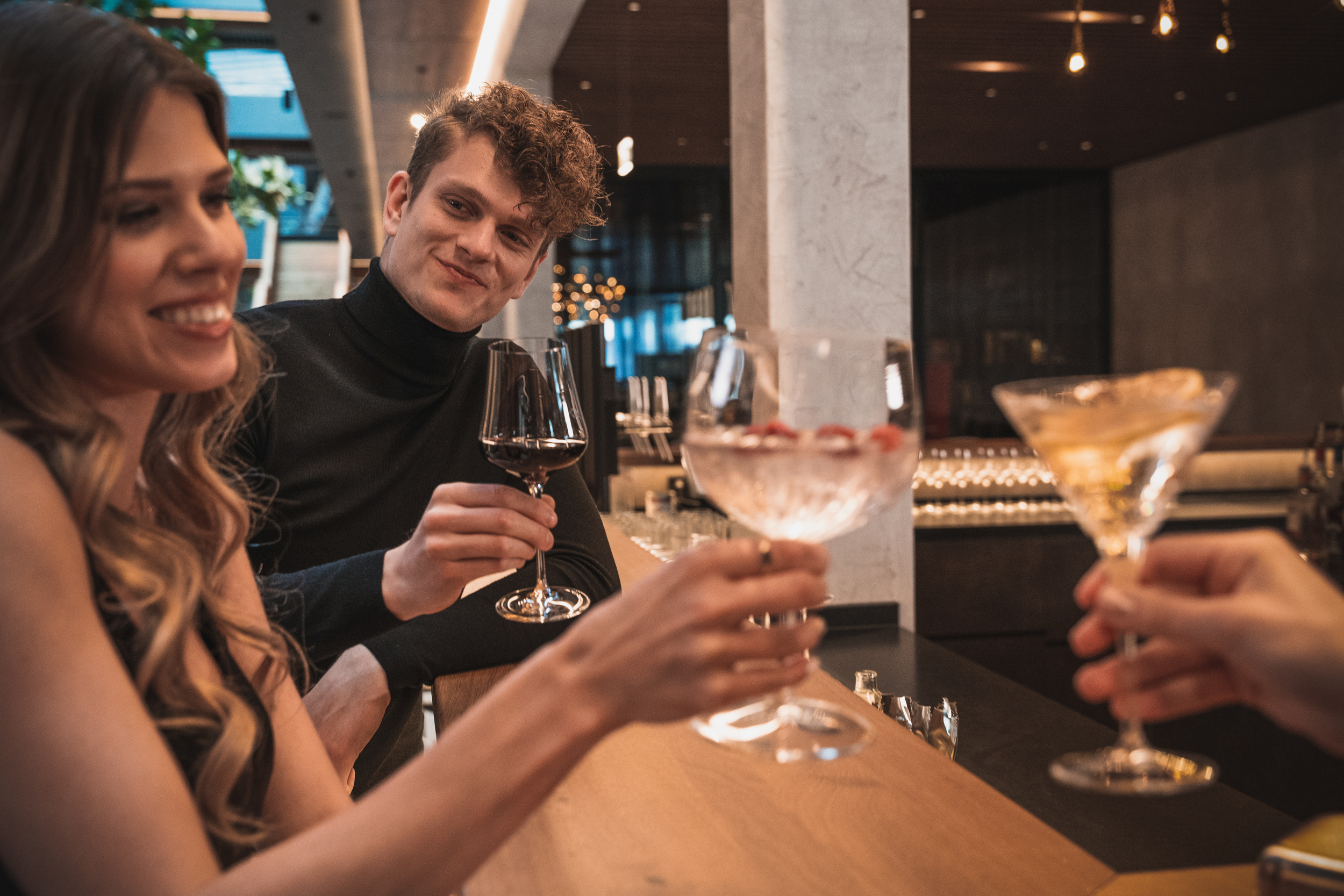 Three people are sitting at a bar, smiling and clinking glasses filled with cocktails and wine.
