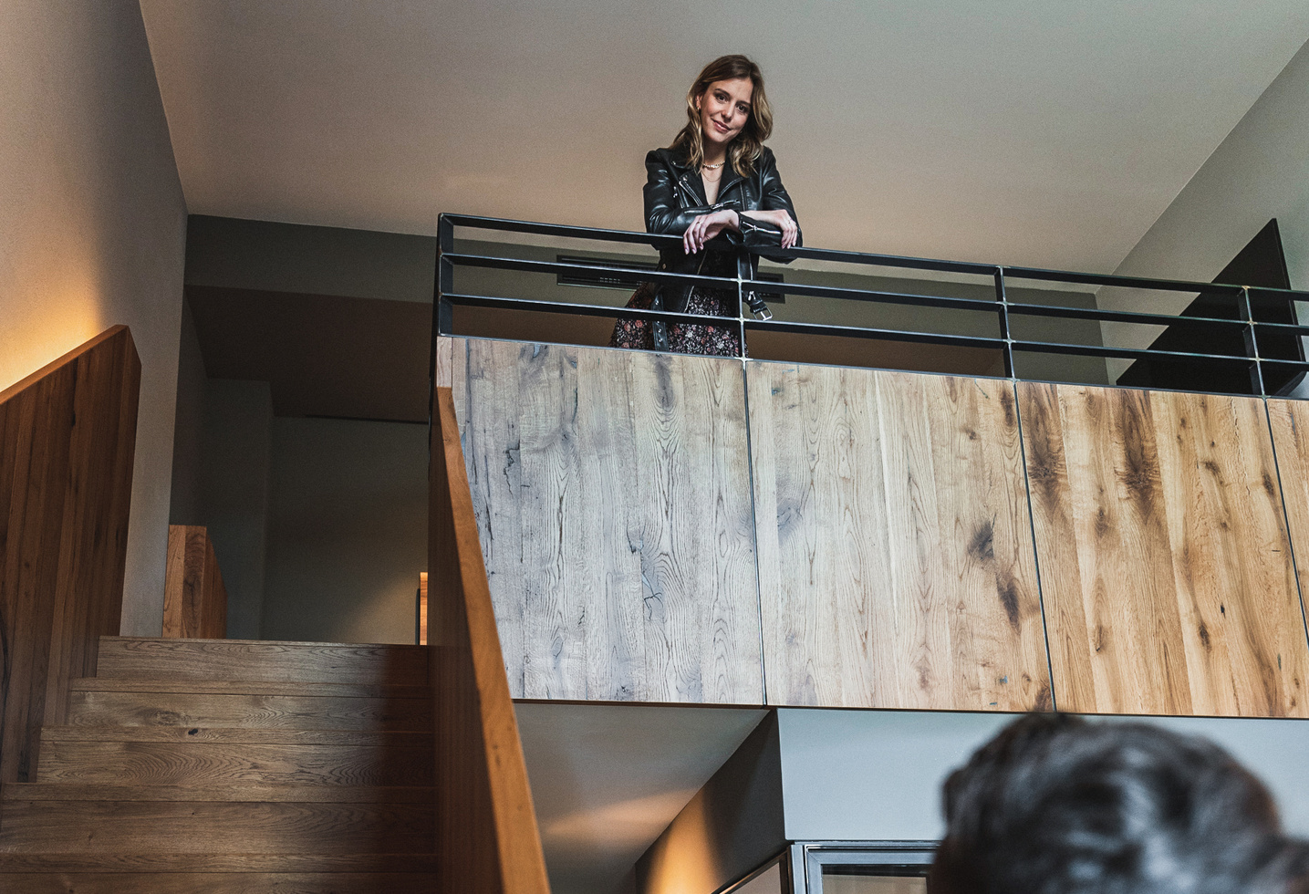 A woman in a black jacket stands on a covered balcony, looks down and smiles.