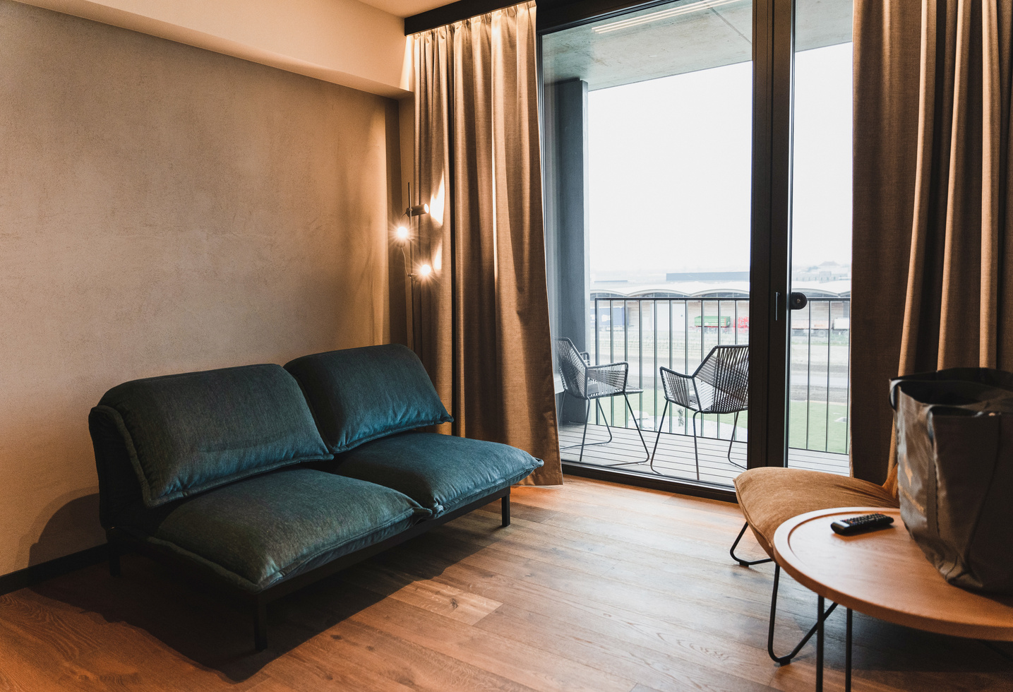 Modern living room with teal-colored sofa, access to the balcony and outdoor chairs, with soft natural light.