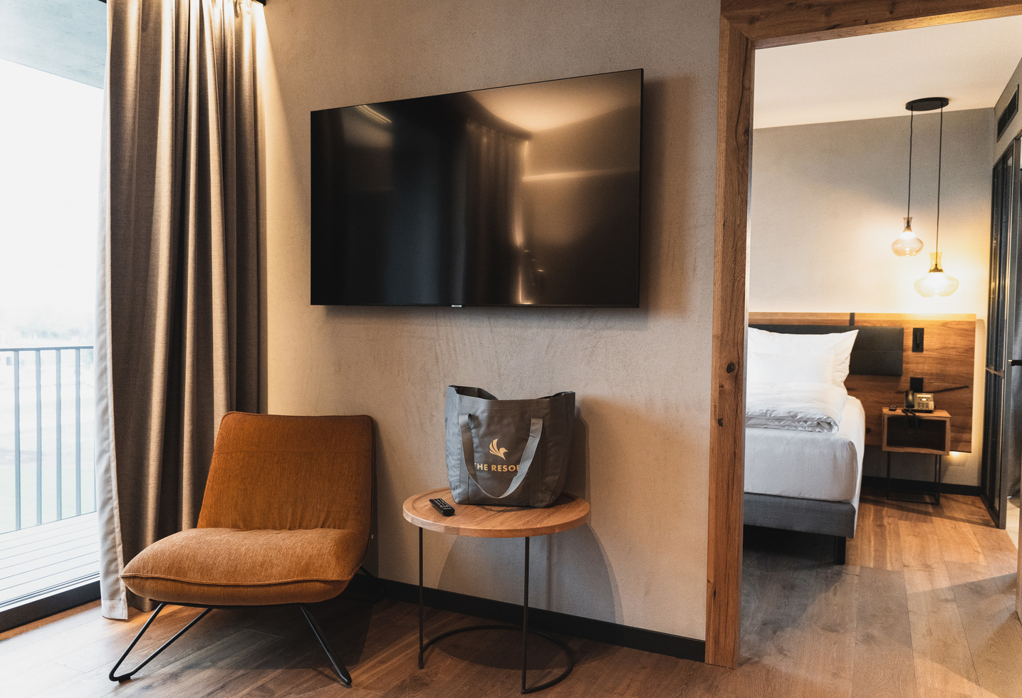 Modern hotel room with a TV, a chair, a small round table and a view into a bedroom with a made-up bed.