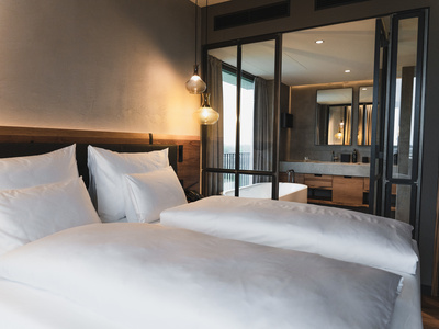 Ein modernes Hotelzimmer mit zwei sauber gemachten Betten, weißer Bettwäsche und einem Kopfteil aus Holz. Glastüren führen in ein Badezimmer mit Badewanne, großem Spiegel und modernen Armaturen.