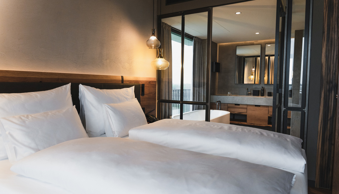 A modern hotel room with two neatly made-up beds, white bed linen and a wooden headboard. Glass doors lead into a bathroom with a bathtub, large mirror and modern fittings.