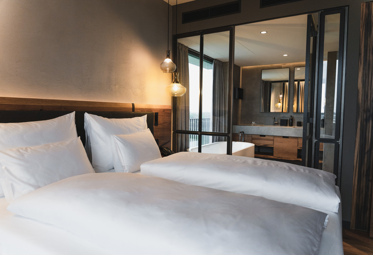 A modern hotel room with two neatly made-up beds, white bed linen and a wooden headboard. Glass doors lead into a bathroom with a bathtub, large mirror and modern fittings.