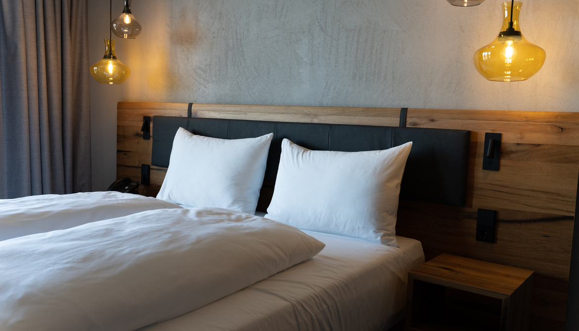 A neatly made bed with white pillows and bedding, a wooden headboard and hanging pendant lights on either side, in front of a gray, textured wall. A small wooden bedside table stands next to the bed.