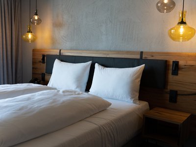 Ein ordentlich gemachtes Bett mit weißen Kissen und Bettzeug, einem hölzernen Kopfteil und hängenden Pendelleuchten an beiden Seiten, vor einer grauen, strukturierten Wand. Ein kleiner hölzerner Nachttisch steht neben dem Bett.