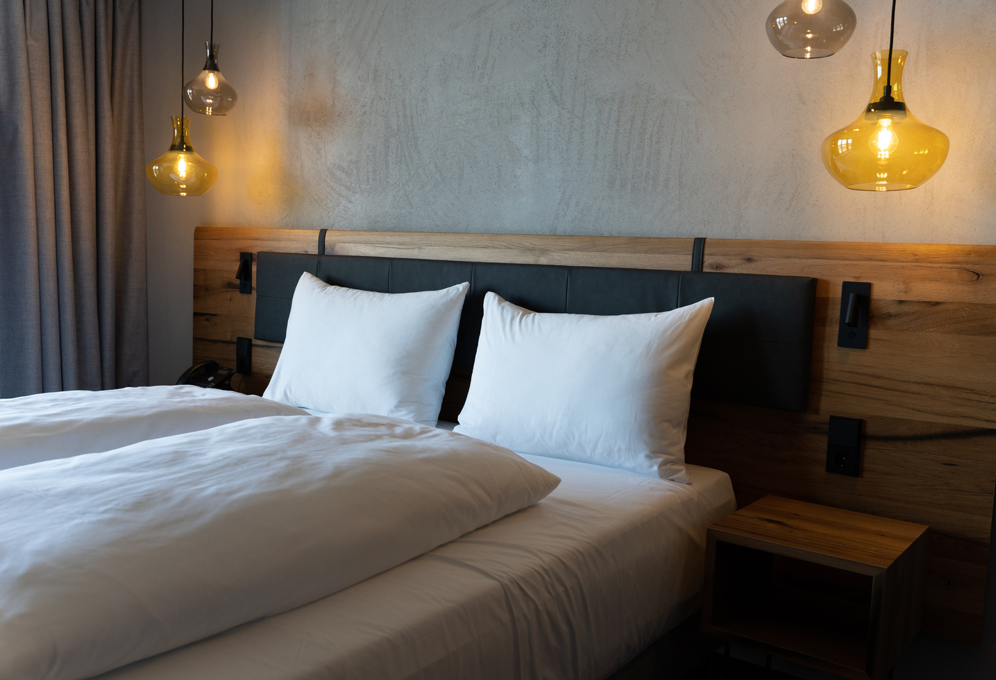 A neatly made bed with white pillows and bedding, a wooden headboard and hanging pendant lights on either side, in front of a gray, textured wall. A small wooden bedside table stands next to the bed.