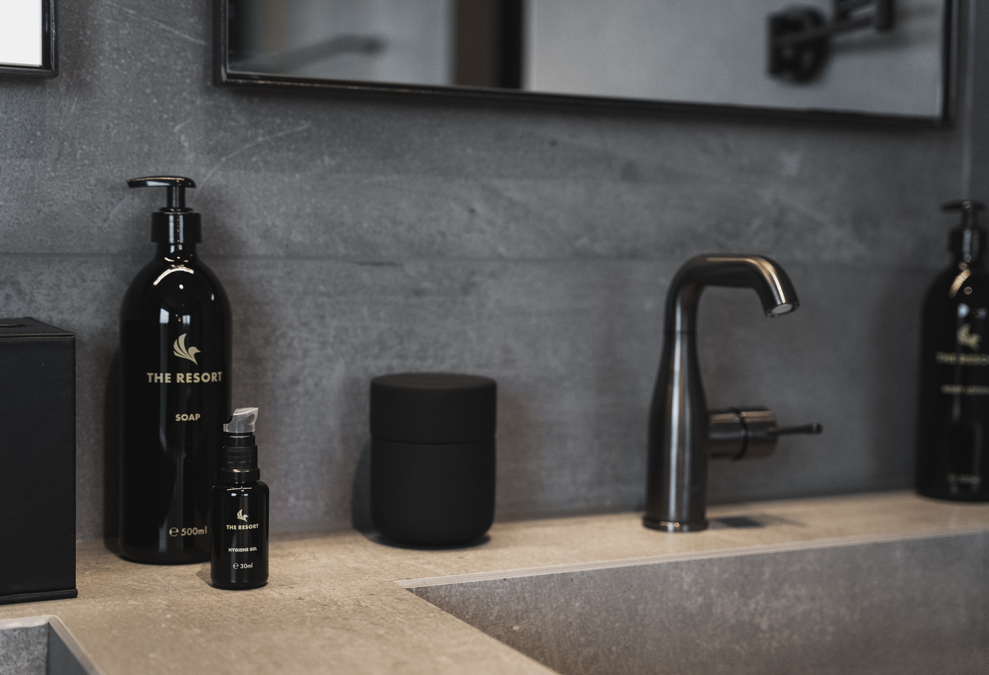 Modern washbasin with black soap dispensers, a black tap and a gray worktop under a mirror.