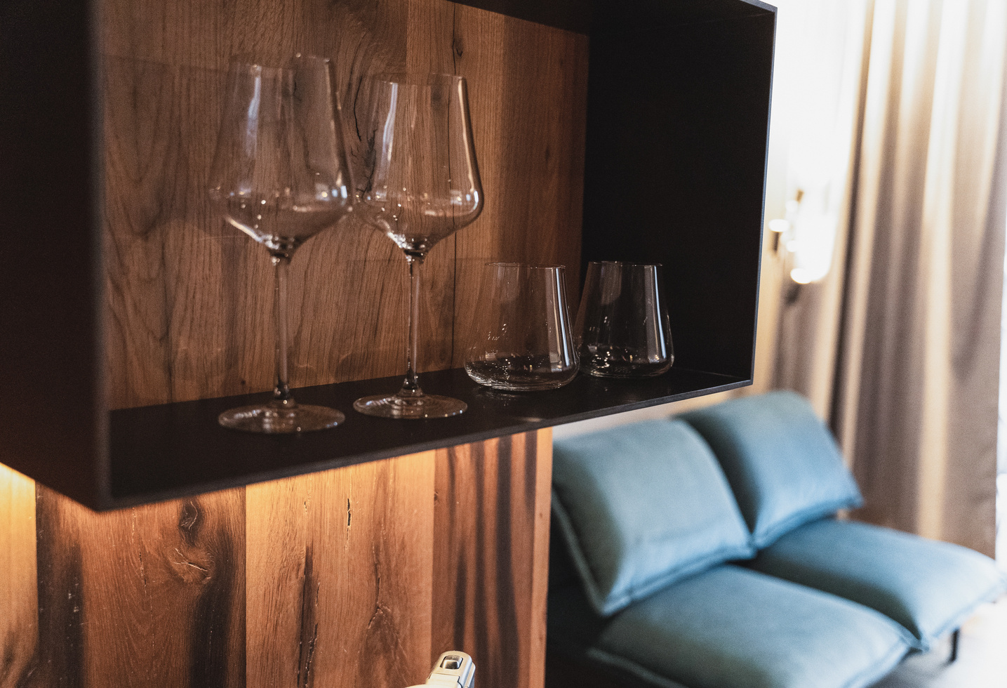 Four empty wine glasses on a wooden shelf with a blue sofa and curtains in the background.
