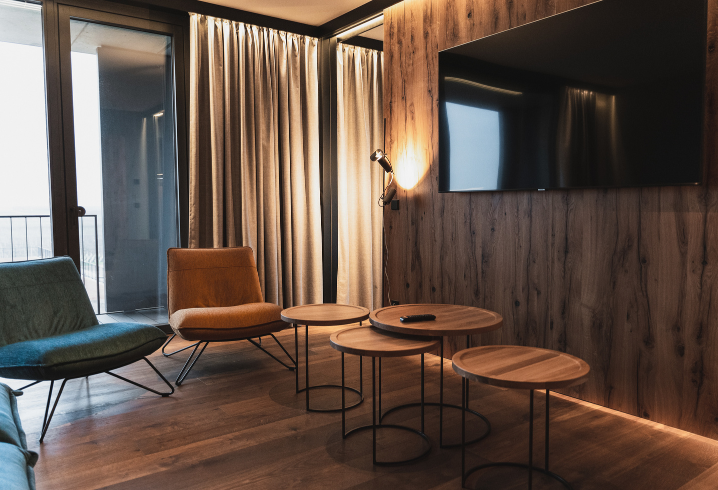 Modern living room with wood paneling, round nested tables, chairs, TV on the wall and floor-to-ceiling curtains.