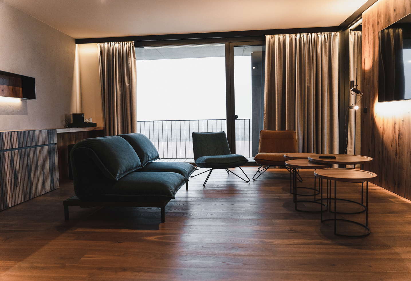 Modern living room with dark sofas, round wooden tables and floor-to-ceiling windows with curtains.