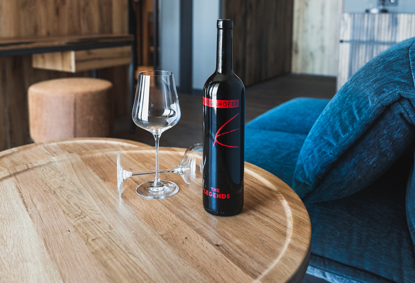 Eine Weinflasche und zwei leere Weingläser auf einem runden Holztisch neben einem blauen Sofa.