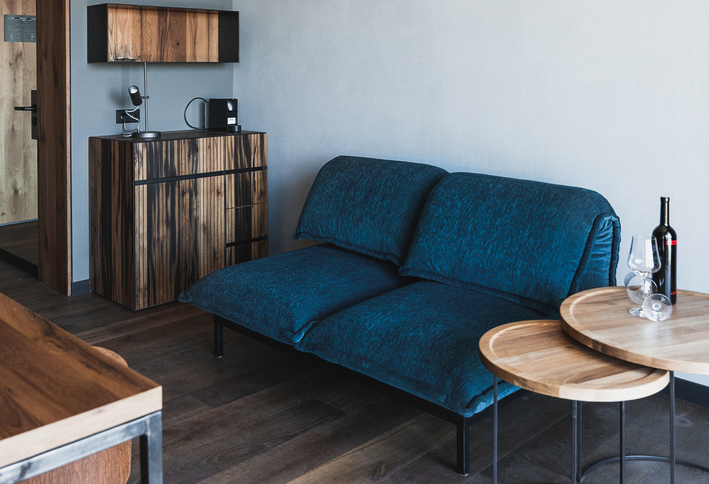 Modernes Wohnzimmer mit einem blauen Sofa, Holztischen und einem Schrank; Wein und Gläser auf dem Tisch.