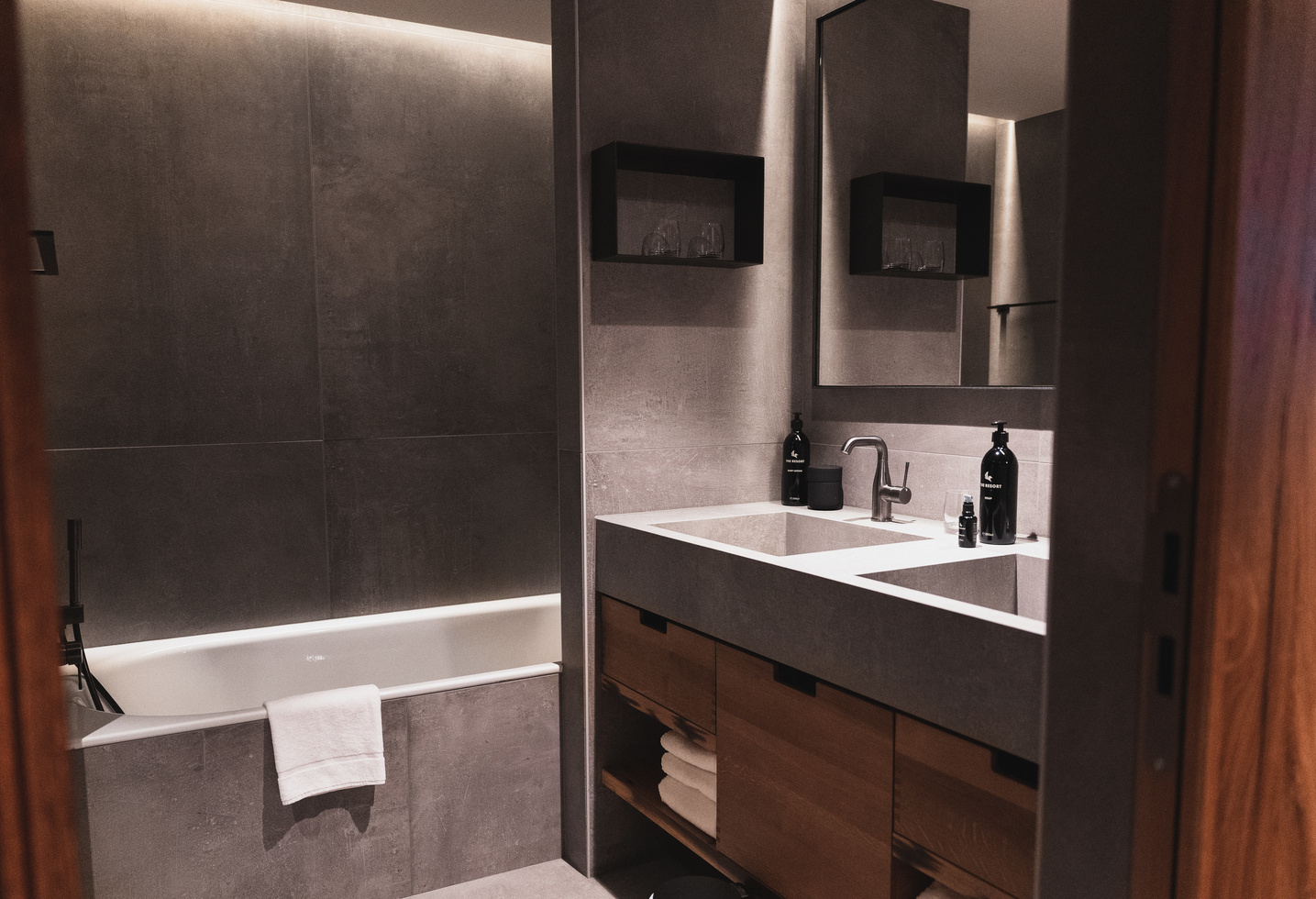 Modern bathroom with double washbasin, wooden cabinets, bathtub and soft lighting; towel hangs on the edge of the bathtub.