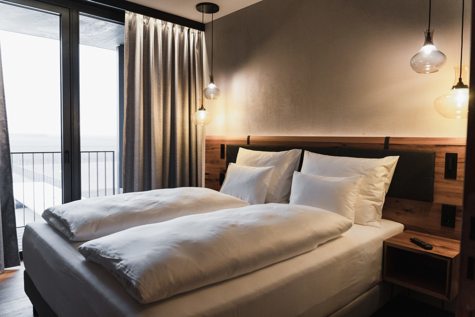 A modern hotel room with a neatly made double bed, white bedding, a wooden headboard, a bedside table, pendant lights, a floor-to-ceiling window, gray curtains and a balcony outside.