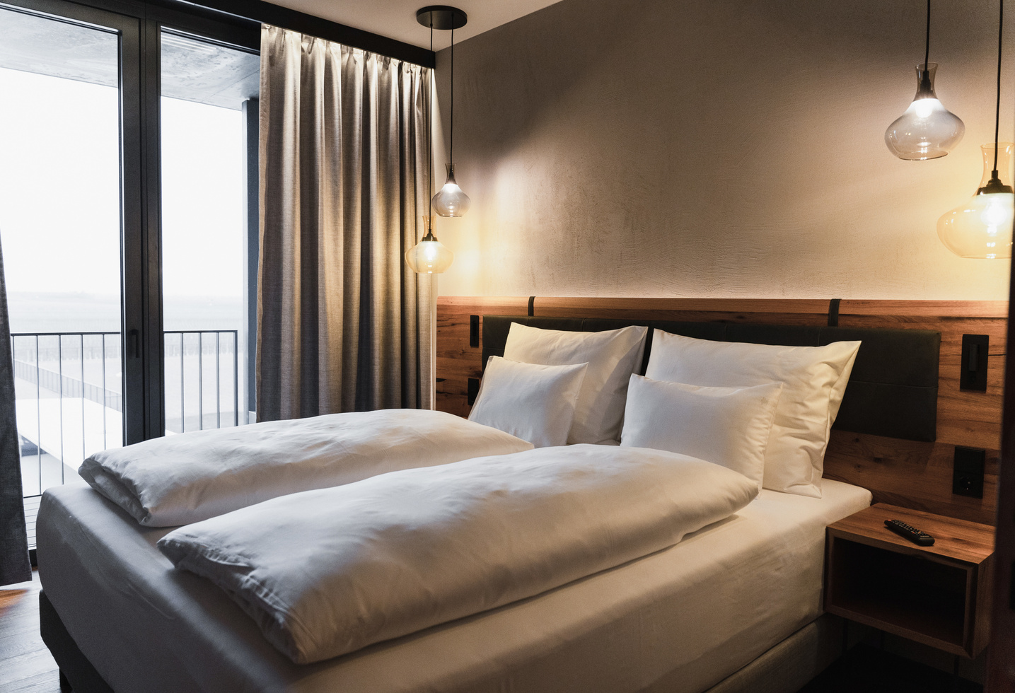 A modern hotel room with a neatly made double bed, white bedding, a wooden headboard, a bedside table, pendant lights, a floor-to-ceiling window, gray curtains and a balcony outside.