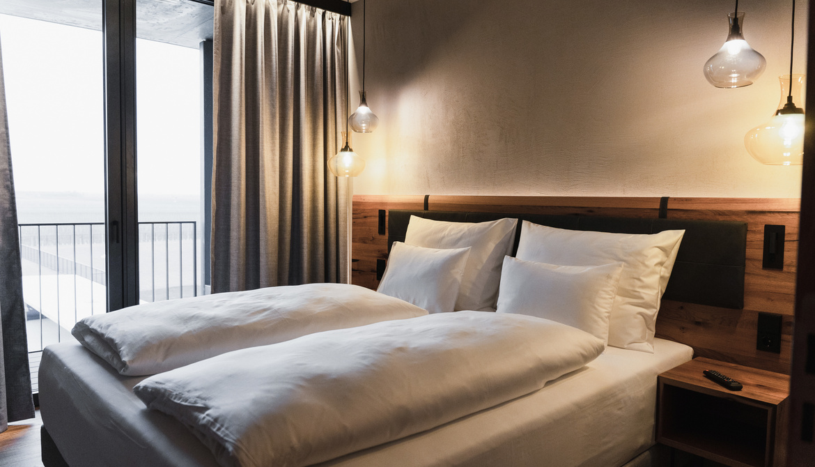 A modern hotel room with a neatly made double bed, white bedding, a wooden headboard, a bedside table, pendant lights, a floor-to-ceiling window, gray curtains and a balcony outside.