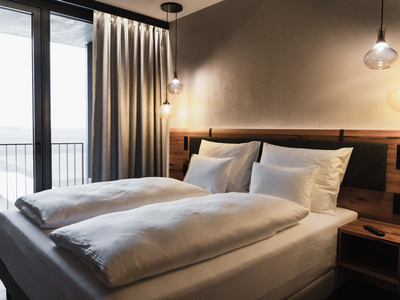 Ein modernes Hotelzimmer mit einem sauber gemachten Doppelbett, weißem Bettzeug, einem hölzernen Kopfteil, einem Nachttisch, Pendelleuchten, einem bodentiefen Fenster, grauen Vorhängen und einem Balkon draußen.