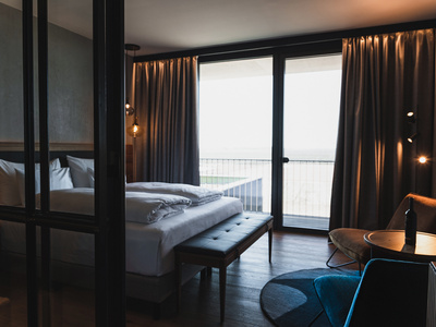 Ein modernes Hotelzimmer mit einem großen Bett, einer Bank am Fußende, bodentiefen Fenstern mit zugezogenen Vorhängen, einem kleinen runden Tisch, Stühlen und sanfter Beleuchtung. Die Fenster öffnen sich zu einem Balkon mit Blick nach draußen.