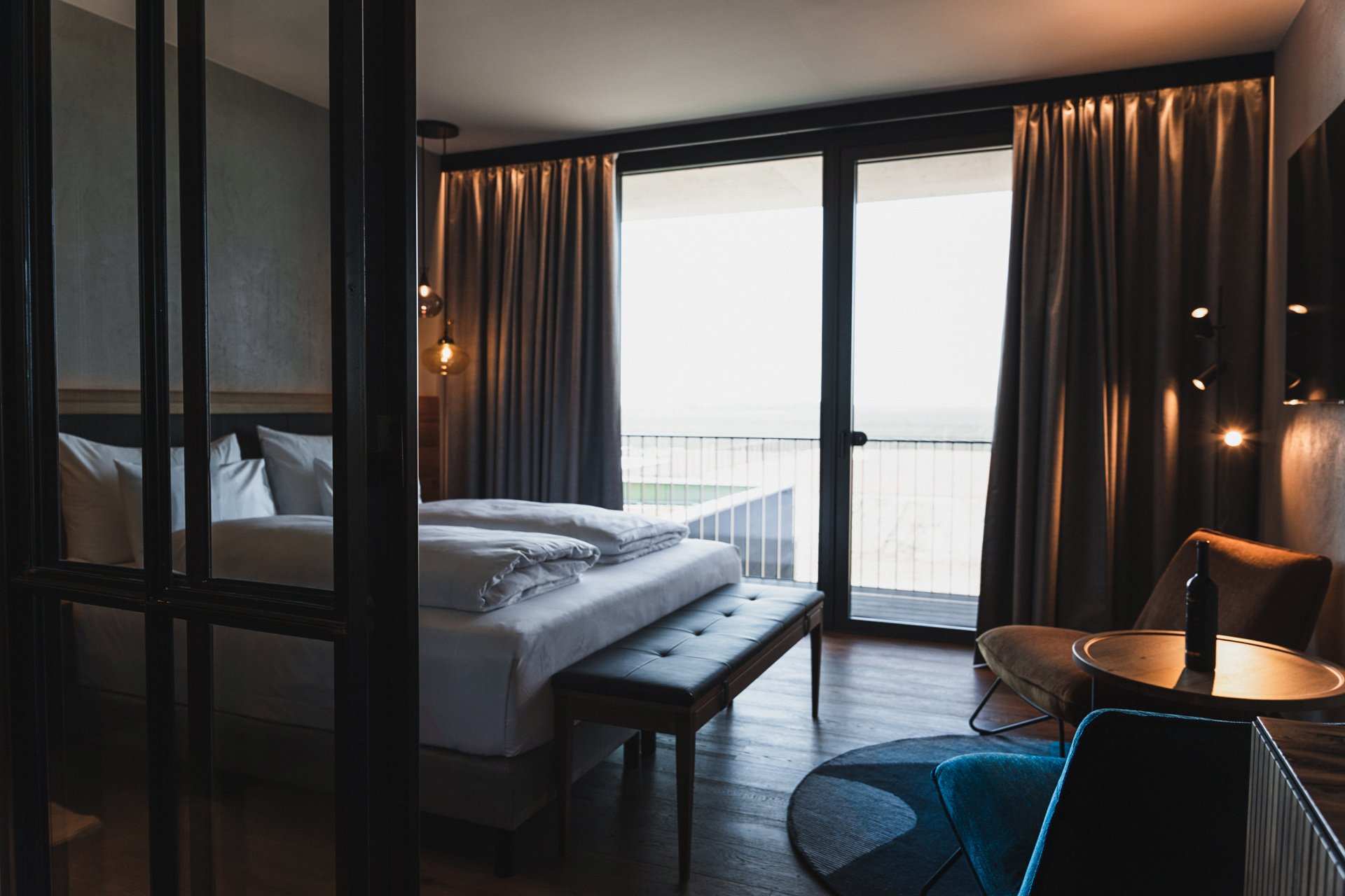 A modern hotel room with a large bed, a bench at the foot of the bed, floor-to-ceiling windows with drawn curtains, a small round table, chairs and soft lighting. The windows open onto a balcony with a view outside.