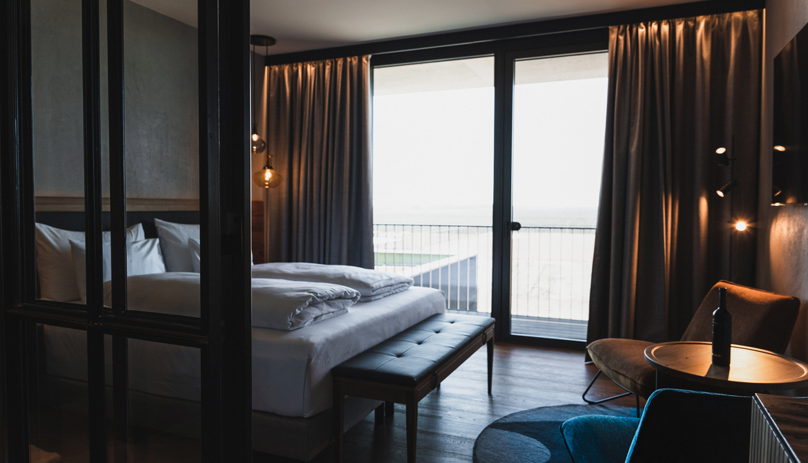 A modern hotel room with a large bed, a bench at the foot of the bed, floor-to-ceiling windows with drawn curtains, a small round table, chairs and soft lighting. The windows open onto a balcony with a view outside.