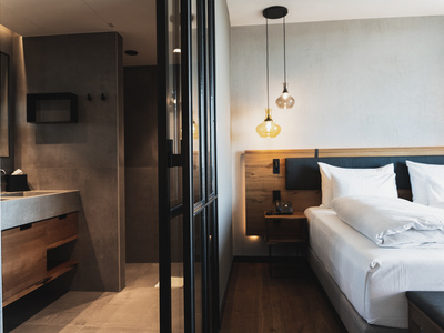 Modernes Hotelzimmer mit einem sauber gemachten Doppelbett, hölzernen Akzenten, Hängelampen und einem offenen Blick auf das angrenzende Badezimmer mit schlichten, minimalistischen Armaturen und einer Arbeitsplatte aus Beton.