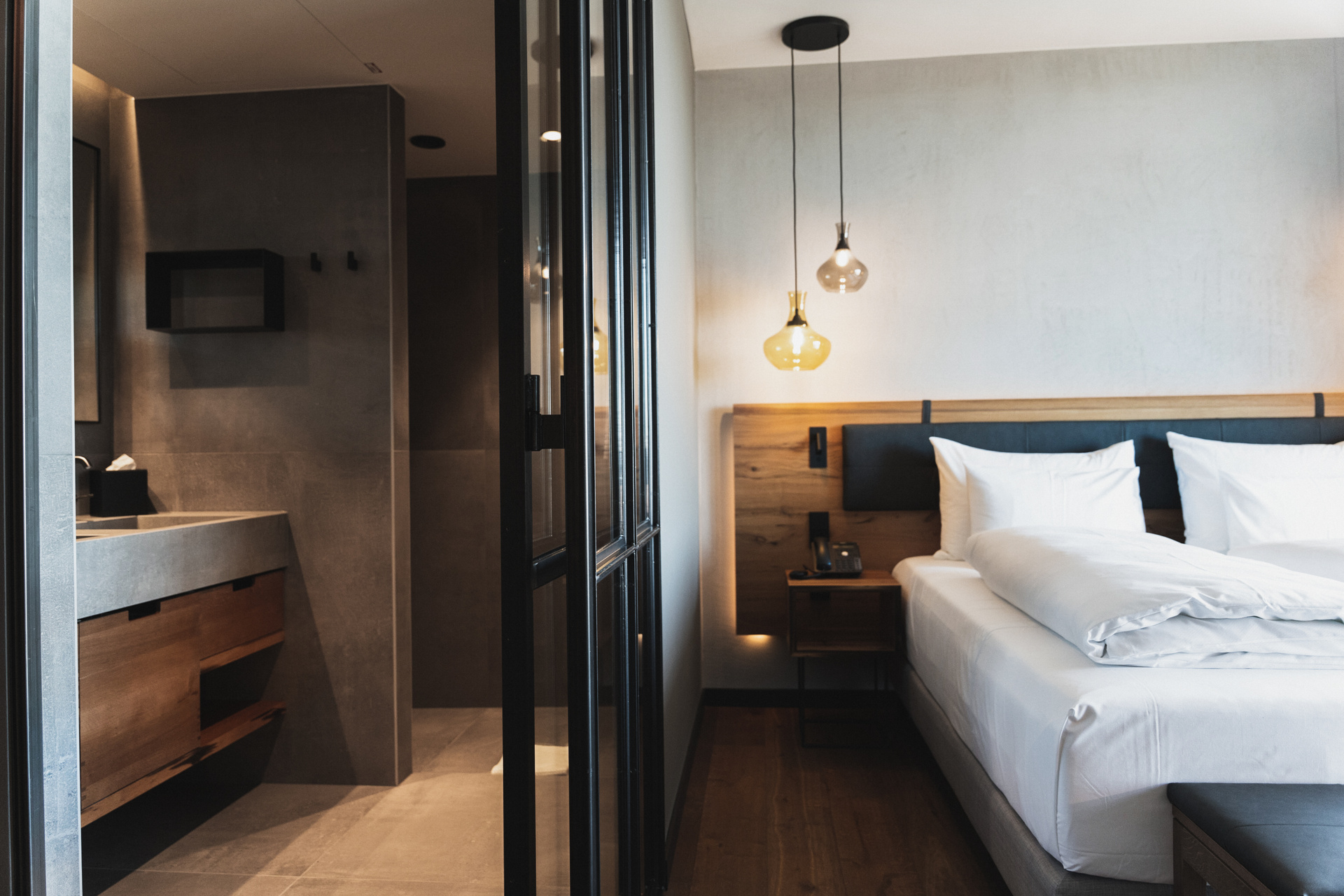 Modern hotel room with a neatly made double bed, wooden accents, hanging lamps and an open view of the adjoining bathroom with sleek, minimalist fixtures and a concrete countertop.
