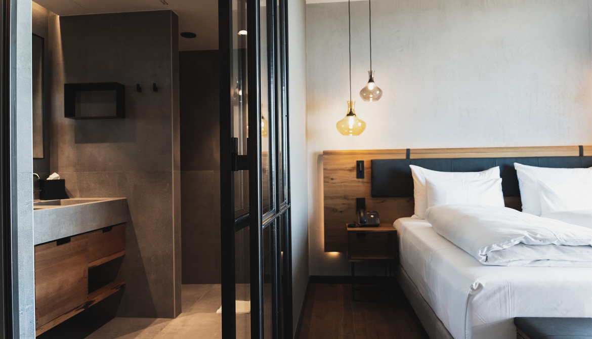 Modern hotel room with a neatly made double bed, wooden accents, hanging lamps and an open view of the adjoining bathroom with sleek, minimalist fixtures and a concrete countertop.