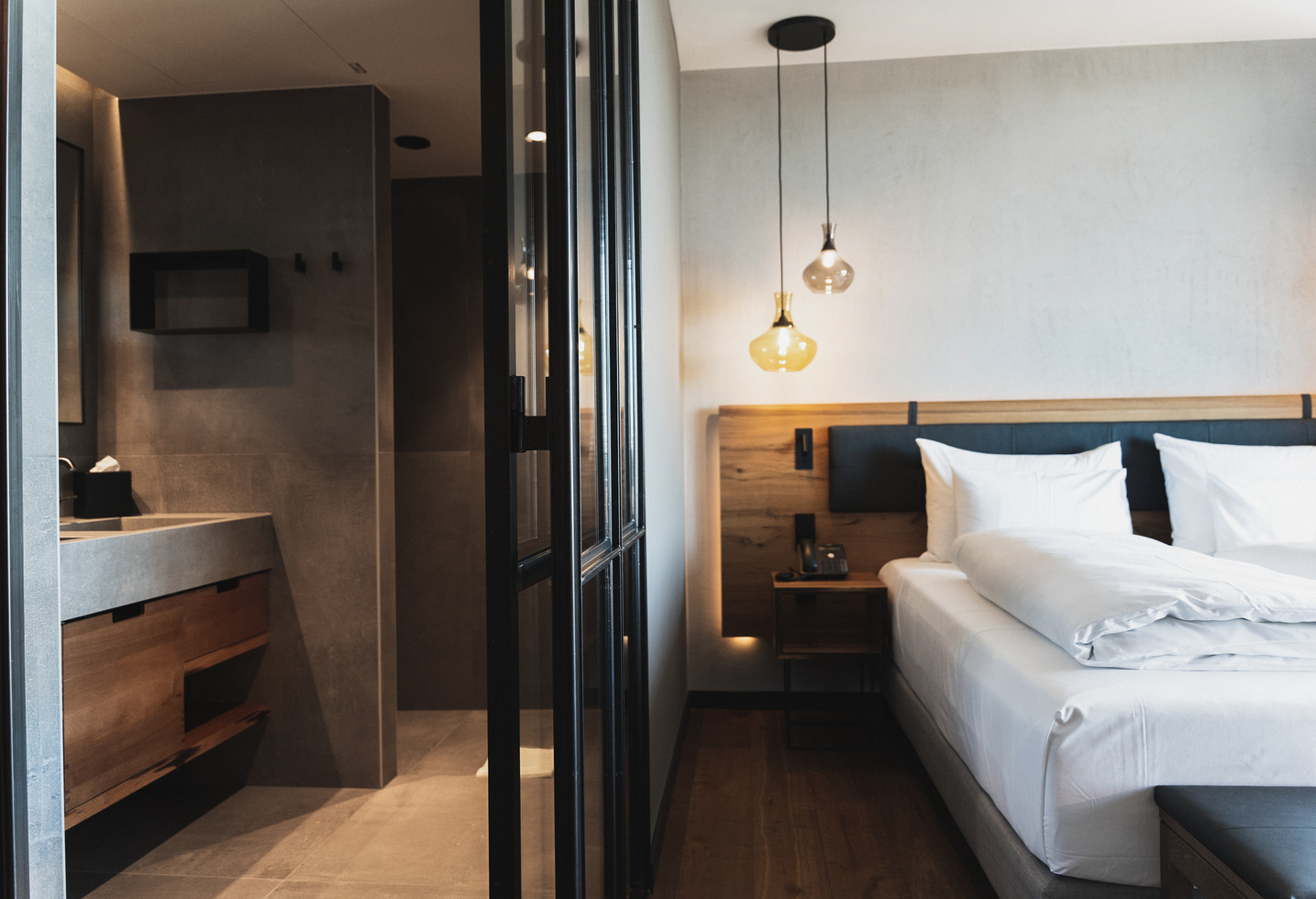 Modern hotel room with a neatly made double bed, wooden accents, hanging lamps and an open view of the adjoining bathroom with sleek, minimalist fixtures and a concrete countertop.