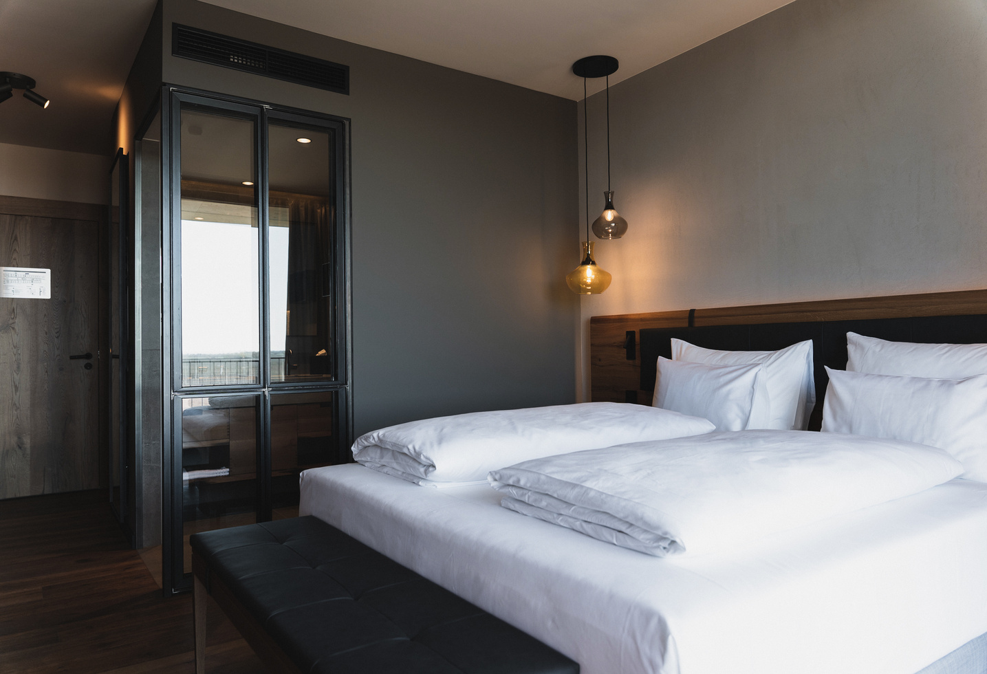 Modern hotel room with double bed, white bed linen, shower with glass walls and warm lighting.