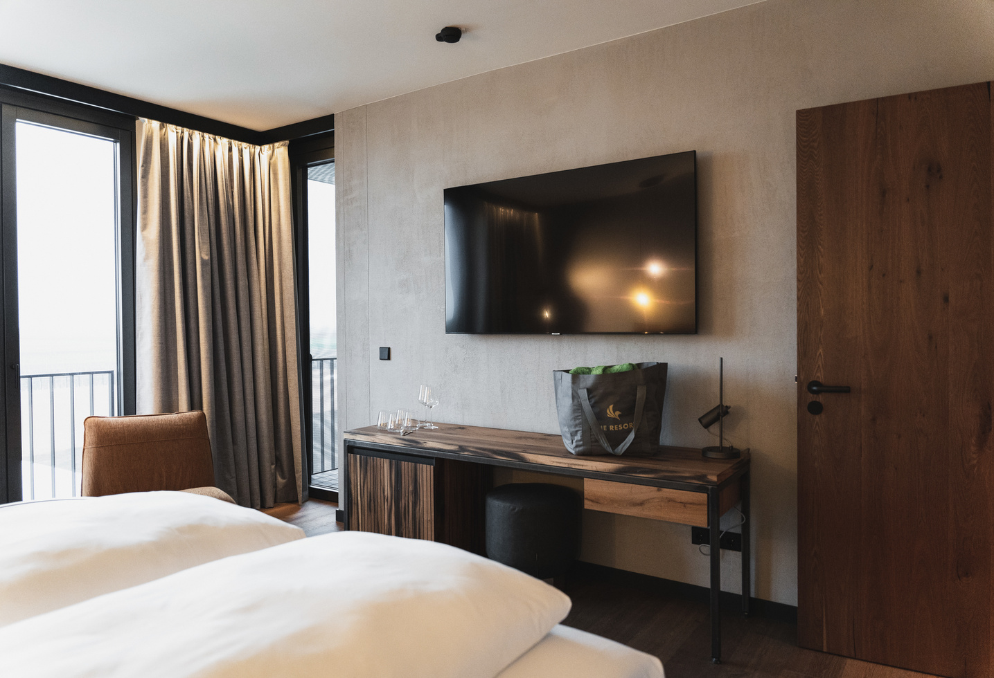 Modern hotel room with a bed, a wall-mounted TV, a desk, a chair and a large window with curtains.