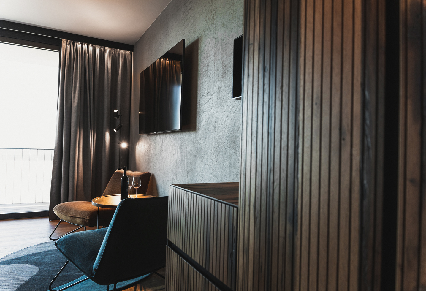 Modern hotel room with wooden furniture, a wall-mounted TV, two chairs, a small table and large window curtains.