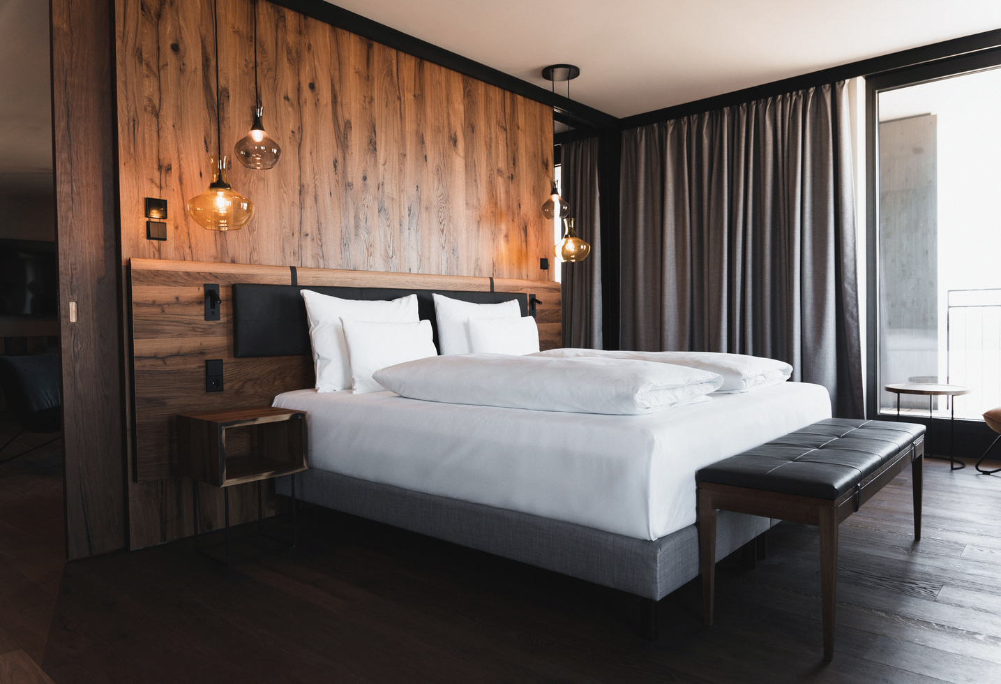 Modern hotel room with large bed, wooden accents, hanging lamps and floor-to-ceiling curtains.