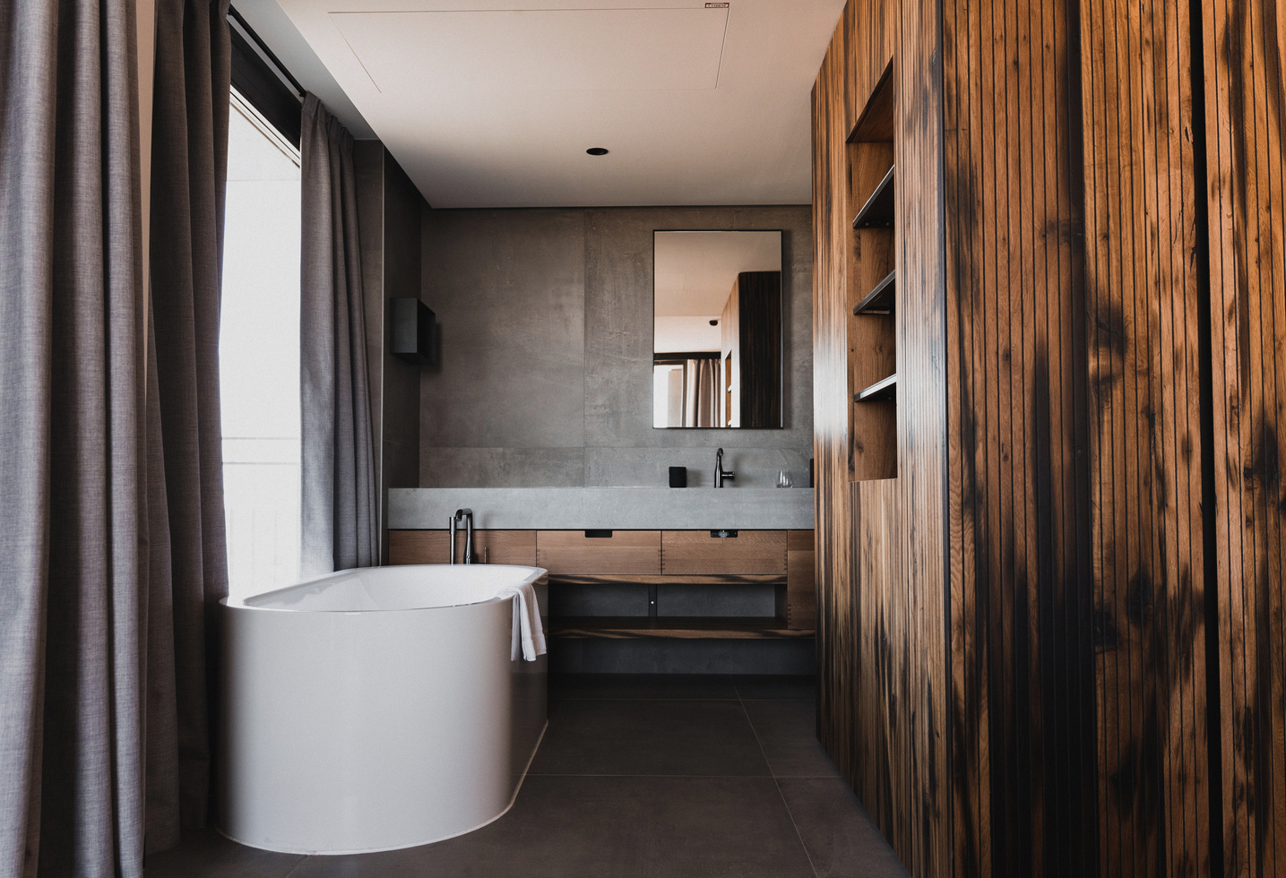 Modern bathroom with a white bathtub, wooden walls, gray curtains and a large mirror above the sink.