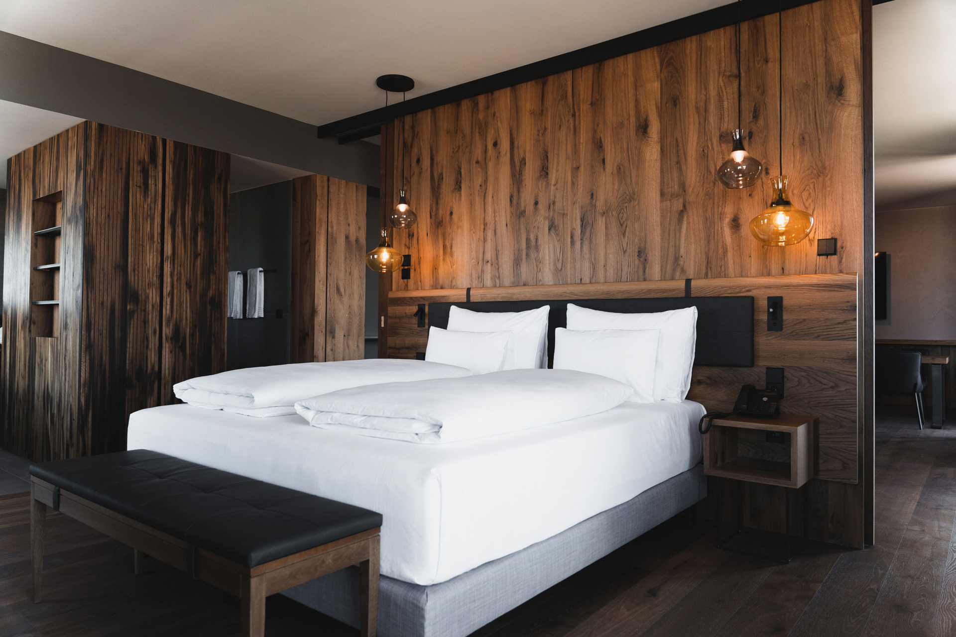 Modern hotel room with large bed, white bed linen, wood-paneled walls and hanging lamps.
