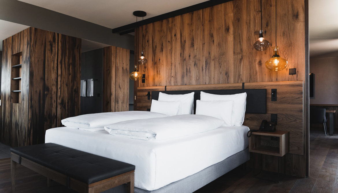 Modern hotel room with large bed, white bed linen, wood-paneled walls and hanging lamps.