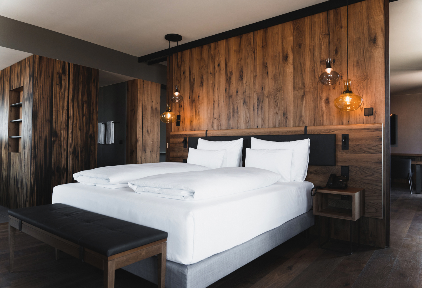 Modern hotel room with large bed, white bed linen, wood-paneled walls and hanging lamps.