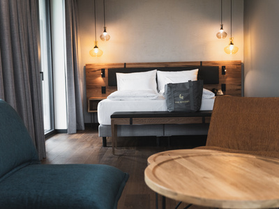 Ein modernes Hotelzimmer mit einem großen Bett, weißem Bettzeug, einem hölzernen Kopfteil, hängenden Pendelleuchten, einem grauen Sessel, einem Holztisch und einer Tragetasche auf dem Bett. Licht strömt durch ein Fenster mit grauen Vorhängen.