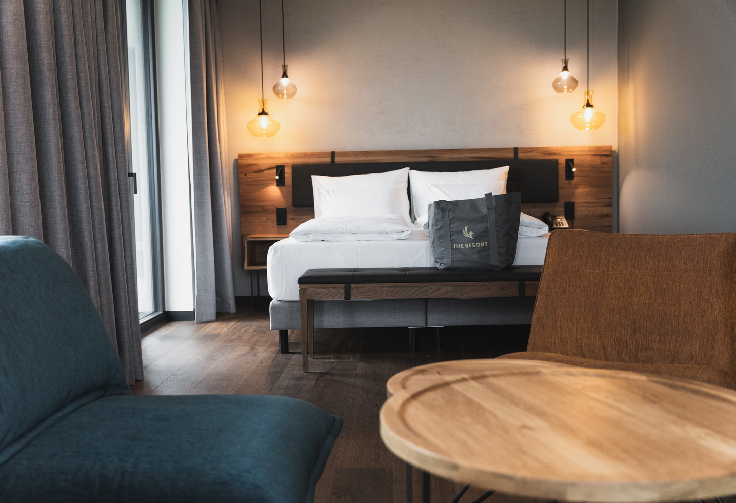 A modern hotel room with a large bed, white bedding, a wooden headboard, hanging pendant lights, a gray armchair, a wooden table and a tote bag on the bed. Light streams in through a window with gray curtains.