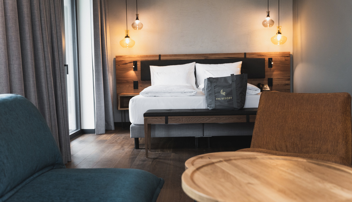 A modern hotel room with a large bed, white bedding, a wooden headboard, hanging pendant lights, a gray armchair, a wooden table and a tote bag on the bed. Light streams in through a window with gray curtains.